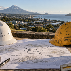 Two hard hats labelled with policy and delivery themes sit on plans overlooking New Plymouth and Mount Taranaki, illustrating central and local government working toward better outcomes from different vantage points.